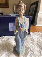 Full view of Lladro figurine showing child in blue overalls holding flowers, standing on base, with original box in background.