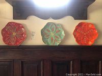 Photo showing set of three colorful South African pottery plates displayed side by side showcasing glossy hand-painted floral and leaf motifs in red, turquoise, and orange colors.