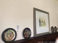 Four decorative plates placed on a dark wooden surface against a wall, one framed art piece to the right