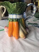 Front and side view of ceramic vegetable and bow motif pitcher with detailed carrots, corn, and turnips beneath a leafy green top and blue-and-white striped handle.