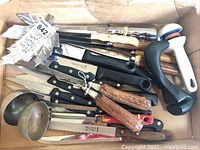 Top view of a box with assorted kitchen utensils including ladles, steak cutters, carving knives, and other kitchen tools with various handle types including wooden and plastic.
