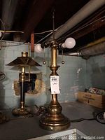 Photo of a single brass table lamp with a solid round base and two light sockets with bulbs, no shade present. Situated in a basement with another lamp and items in the background.