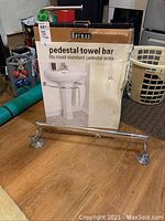Pedestal towel bar in its original box, displayed with the towel bar on the floor showing mounting ends and the box's photo of the towel bar on a pedestal sink.