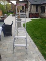 Front view of Lite aluminum step ladder fully extended in outdoor setting on paved tile ground with patio furniture and garden in background.