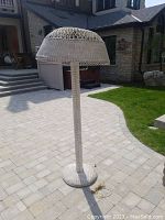 Full view of the tall white wicker floor lamp standing upright on a paved ground outdoors.