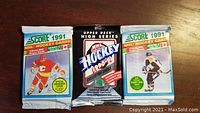 Three unopened packs of hockey cards: one Upper Deck High Series 1990-91 in silver packaging, two Score 1991 NHL Hockey cards in blue packaging shown on wooden surface.