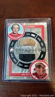 Front of trading card showing circular window with fabric pieces from Gerry Cheevers' goalie pad and Bobby Hull's pants along with their portraits and player career years.