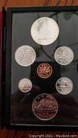 1976 Canadian double dollar coin set displayed in black case with visible damage to case. Contains multiple Canadian coins of various denominations and designs including parliament building, caribou, and maple leaf.