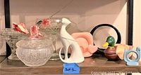 Full view of shelf displaying various mid-century modern decorative items and glassware including a white ceramic bird, pink shell dish, wooden duck figurine, black circular frame, and cut glass candy dish.
