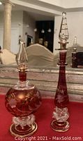 Photo of two hand-blown etched perfume bottles placed on a mirrored surface with reflection visible. One round bottle and one tall slender bottle.