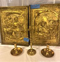 Front view of two brass wall arts with detailed embossed scenes set on wooden frames and three solid brass candle holders placed in front.