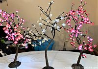 Three illuminated flower tree lamps on a table, showing pink and white lighting flowers with brown bases and trunks.