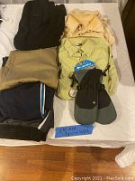 Front view of lot showing clothing neatly folded on bed including black sweater, beige and green shirts, khaki pants, navy shorts, and mittens