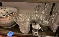 Shelf with large silver bowl holding glass goblets, clear cut glass pitcher, glass bowl with metal stand holding glass stirrers, clear glass container with metal handles, and silver stovetop coffee maker.