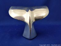 Front view of the shiny aluminum whale tail sculpture on blue background, showing reflective surface and curved design.