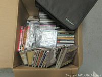 Photo showing the lot in a cardboard box including rows of CDs and DVDs and a black soft case