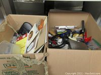 Two cardboard boxes filled with assorted sundry items including books, kitchen utensils, and miscellaneous household goods showing overall lot contents.