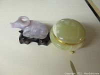 Carved lavender quartz ox statue on a dark ox-shaped wooden stand next to a green onyx dresser box with brass details on top of a surface.