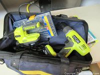 Open fabric tool bag containing the Ryobi drill/driver, charger, one battery, and accessory kit