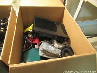 Box with a black Blu-ray player, tangled cables, a small zippered pouch, headphones, and other small electronics