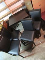 Photo showing four dark wood framed dining chairs with black padded seats and backs arranged around dark square table in storage area.