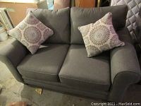 Front view of grey upholstered loveseat with two patterned throw pillows on concrete floor