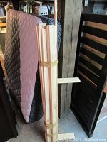 Photo showing queen size mattress with a quilted pattern and bundle of wooden frame slats attached with rubber bands next to a dark wood slatted headboard.