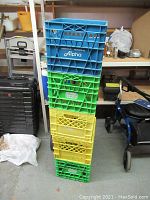 Stack of 5 plastic milk crates in blue, green, and yellow colors showing logos Alpha and Island Farms in used condition.
