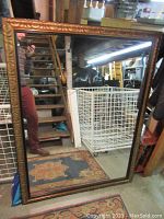 Full front photo of large rectangular mirror with detailed carved gold frame, reflecting surrounding storage items and stairs. Shows overall size and style.