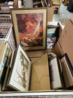 Photo showing multiple framed prints including a sailboat print, bird illustration, and others in varied frames stacked in a box and leaning back.