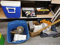 Blue plastic cooler on shelf and two blue bins filled with assorted items including vintage cameras, glassware, and miscellaneous objects on floor around them.