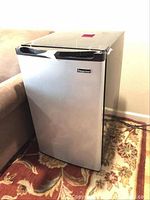 Closed view of silver Magic Chef mini fridge with black trim, placed on patterned rug next to sofa.