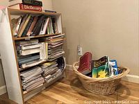 Full view of a bookshelf with a mixture of hardcover and paperback books, some stacked horizontally, some vertically, and a basket with additional books on the hardwood floor beside it.