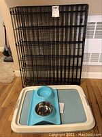 Full view of folded black metal kennel frame with plastic tray and blue feeding dish in front.