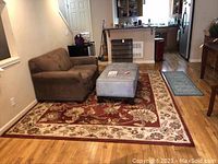 View of larger red floral rug with cream border, loveseat and ottoman placed on top, showing condition and size in room setting.