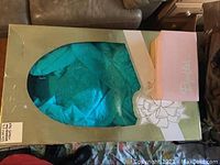 Wedding dress in original box with green frame and a small pink box attached, dress is teal with lace details visible through the box window.