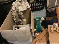 Box with assorted knickknacks including a green ceramic vase inside a wooden box with Japanese characters around it, with some packaging materials.