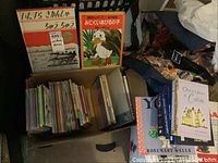 Photo of a box filled with various children's books, mostly in Japanese with colorful illustrated covers. Two larger books are prominently displayed on top, one featuring a duck character (Disney) and a Japanese title.