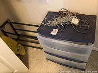 Plastic three drawer storage unit with blue top and handles, black metal shoe rack, string Christmas lights and extension cord tangled on top.