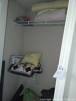 Closet or storage area with several blankets, pillows, and linens stored on wire shelving and some on the floor. Visible multicolored patchwork blanket and a yellow pillow.