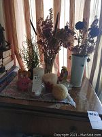 Photo showing home decor items including vases with dried flowers, candles, candle holders, pig-shaped object, fairy figurine, all arranged on a doily.