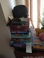 Stack of books topped with a black binocular case and red basket with charging cords and adapters, visible Monopoly Disney Pixar board game box at bottom