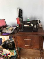 Vintage White Rotary sewing machine in dark wood cabinet with drawers, black cast iron machine with metallic accents, spool of green thread on top, sewing attachments visible around.
