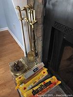 Brass-colored duck head fireplace tools standing by stone fireplace with three yellow boxes of Duraflame fire logs and brass spittoon with kindling sticks on floor.