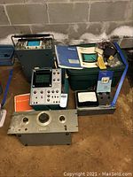 Overview of oscilloscope, multiple audio oscillators, and manuals on floor around plastic bin and concrete wall