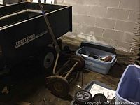 Partial side and front view of manual reel mower with blue parts tub nearby in basement setting