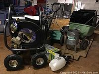 Photo showing metal hose reel cart with hose, seed spreaders, sand blaster, buckets, sprayer, and soaker hose arranged on basement floor.
