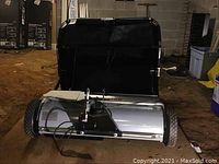 Front view of the lawn sweeper showing the metal brush housing, wheels, and black collection bag behind.