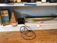 Wide view of shelf with Seth Thomas metronome in box, Franz electric metronome with dial, long black model sailboat body, miscellaneous parts and power cords on wooden floor.
