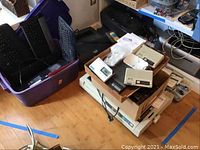 Overview of multiple electronic items including keyboards in purple tub and stacked printers and devices on floor